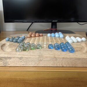 May include: A wooden game board with a grid pattern and a raised edge. The board is filled with glass marbles in various colors, including blue, green, and brown. The marbles are arranged in a pattern on the board.