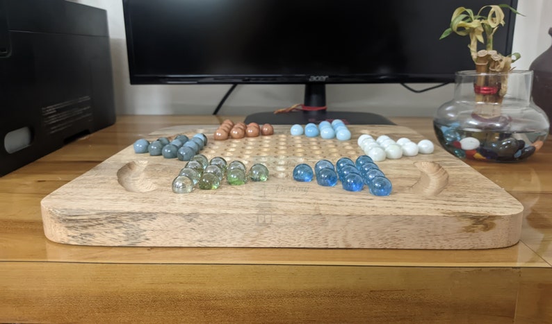 May include: A wooden game board with a grid pattern and a raised edge. The board is filled with glass marbles in various colors, including blue, green, and brown. The marbles are arranged in a pattern on the board.