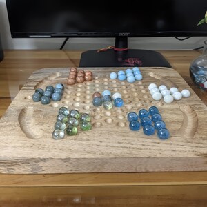 May include: A wooden game board with a grid of holes and a variety of colored marbles. The marbles are arranged in groups of different colors, including blue, green, brown, and white.