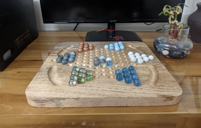 May include: A wooden game board with a grid of holes and a variety of colored marbles. The marbles are arranged in groups of different colors, including blue, green, brown, and white.