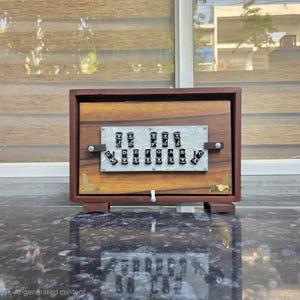 Könnte beinhalten: Ein rechteckiges hölzernes Musikinstrument mit einem dunkelbraunen Rahmen und einer hellen Holzmaserung auf der Vorderseite. Eine zentrale weiße, texturierte Platte zeigt zwei Reihen schwarzer Schalter, die mit den Noten C, D, E, F, G, A, B und C beschriftet sind.