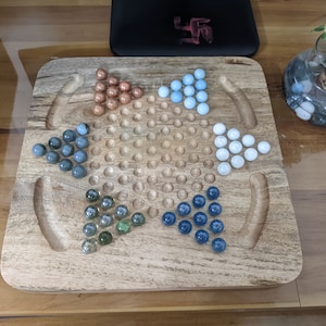 May include: A wooden game board with a star-shaped pattern in the center. The board has four sections, each with a different color of marbles: blue, green, brown, and white.