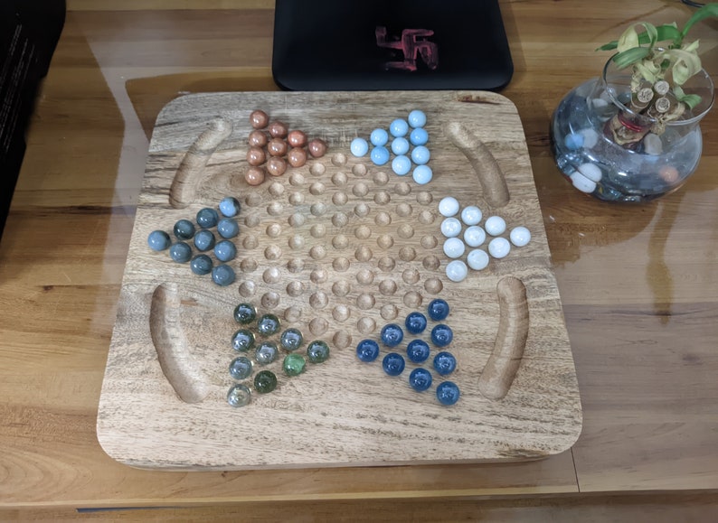 May include: A wooden game board with a star-shaped pattern in the center. The board has four sections, each with a different color of marbles: blue, green, brown, and white.