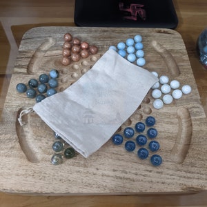 May include: A wooden game board with a white cloth bag and glass marbles in blue, white, and brown colors. The board has a carved design and is made of wood.