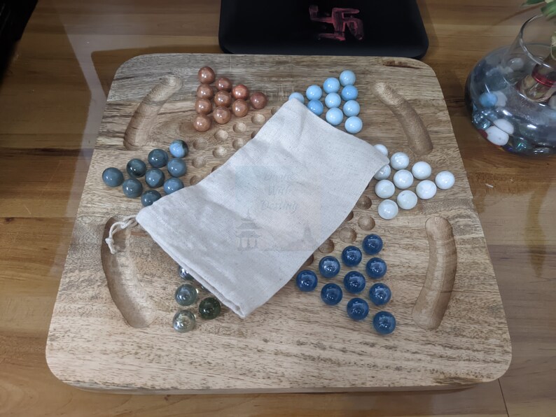 May include: A wooden game board with a white cloth bag and glass marbles in blue, white, and brown colors. The board has a carved design and is made of wood.