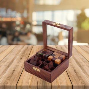Puede incluir: Una caja de madera de color marrón oscuro con tapa abatible y parte superior de cristal transparente, que cuenta con un cierre de metal dorado. En su interior hay varias piezas de rompecabezas de madera, incluyendo un cubo y formas geométricas.