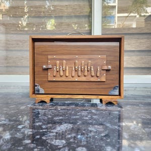 Puede incluir: Un instrumento musical de madera rectangular, un armonio, con un acabado de veta de madera marrón oscuro. El panel frontal muestra una placa rectangular central con once teclas verticales de madera, detalles metálicos en las esquinas y un asa.