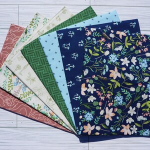 K&ouml;nnte beinhalten: Ein Set aus acht Stoffquadraten mit verschiedenen Mustern, darunter florale, geometrische und Waldmotive. Die Stoffe sind in Blaut&ouml;nen, Gr&uuml;n, Wei&szlig; und Rosa.