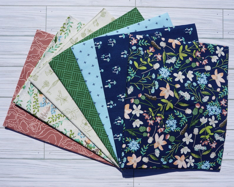 K&ouml;nnte beinhalten: Ein Set aus acht Stoffquadraten mit verschiedenen Mustern, darunter florale, geometrische und Waldmotive. Die Stoffe sind in Blaut&ouml;nen, Gr&uuml;n, Wei&szlig; und Rosa.