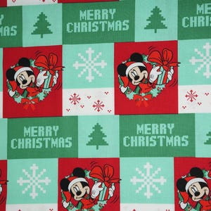 May include: Fabric featuring a repeating pattern of Mickey Mouse wearing a Santa hat and holding a gift, interspersed with pixelated Christmas trees, snowflakes, and the text Merry Christmas in red, white, and green, with a 9-inch ruler visible.