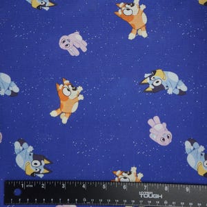 May include: A blue fabric featuring a repeating pattern of cartoon dogs in orange and light blue, interspersed with small light purple figures. A black ruler with white markings is positioned at the bottom, showing a scale in inches.