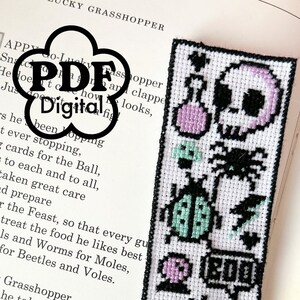 May include: White cross-stitch bookmark with black border and fringe. Features a skull, spider, ladybug, and eye design in black, pink, and green. The word "BOO" is also stitched on the bookmark. The bookmark is on top of an open book.