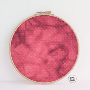 Puede incluir: Un bastidor de bordado redondo de madera que contiene una tela teñida en tonos de rojo y rosa oscuro. La tela tiene un aspecto texturizado, con un efecto marmolado o teñido anudado. El bastidor está sobre un fondo blanco.