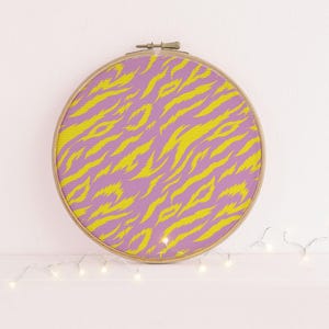 May include: A circular wooden embroidery hoop containing fabric with a vibrant yellow and purple tiger stripe pattern. The fabric is stretched taut within the frame, which features a metal tightening screw at the top centre of the circular hoop.