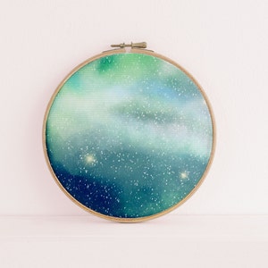 May include: A round, wooden embroidery hoop with a green and blue watercolor painting of a starry night sky. The painting features white stars and a swirling nebula.