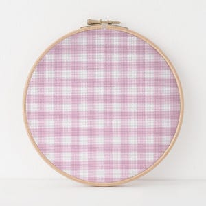 May include: A cross-stitch embroidery hoop featuring a pink and white gingham pattern. The fabric is stretched taut within a round, wooden hoop. The design is a classic, repeating check pattern, perfect for home decor or craft projects.