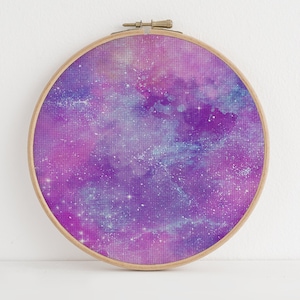 Könnte beinhalten: Ein runder Stickrahmen mit einem Aquarell-Galaxie-Design in Lila und Blau. Das Design zeigt einen wirbelnden Nebel mit weißen Glitzersternen.