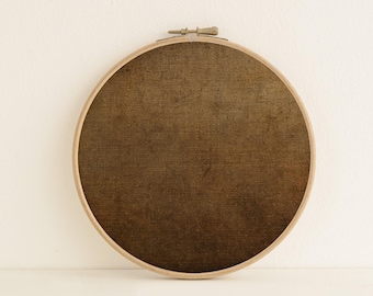 May include: A circular wooden embroidery hoop framing a piece of dark brown, textured fabric. The fabric has a visible woven grain and a mottled, earthy appearance. The hoop features a metal tightening screw at the top center of the frame.
