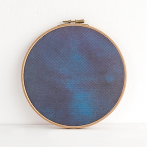 May include: A circular embroidery hoop featuring a blue and navy fabric design. The fabric has a watercolour effect with lighter blue patches. The wooden hoop is secured with a brass clasp.