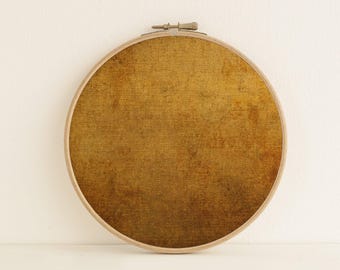 May include: A circular wooden embroidery hoop framing a piece of fabric with a textured, mottled golden-brown surface. The fabric displays a subtle horizontal lined pattern with darker brown patches, secured by a metal tension screw at the top.