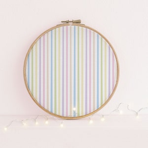 Puede incluir: Un bastidor de bordado redondo con un diseño de tela a rayas pastel. La tela presenta rayas verticales en tonos rosa, azul, amarillo y verde. El bastidor es de madera clara y tiene un cierre metálico.