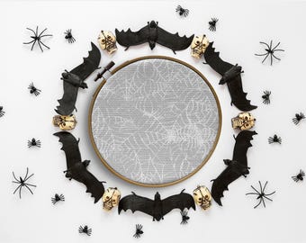 Puede incluir: Un aro de bordado blanco con un diseño de telaraña rodeado de murciélagos negros, calaveras y arañas sobre un fondo blanco.
