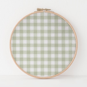 Puede incluir: Un bastidor de bordado redondo con un patrón de cuadros vichy verde claro y blanco. La tela está tensada dentro de un aro de madera natural, creando una pieza decorativa. El diseño de cuadros vichy es un patrón clásico en forma de cuadrícula.