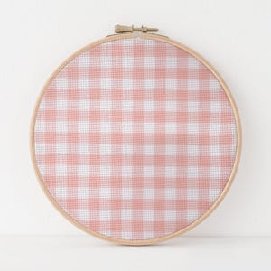May include: A round embroidery hoop with a pink and white gingham pattern. The fabric is stretched taut within a wooden hoop, creating a decorative piece. The gingham design is a classic pattern, often associated with picnics and country themes.