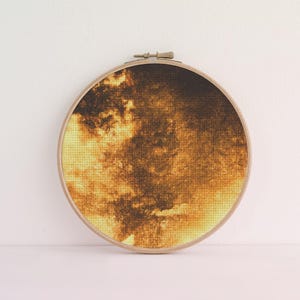 May include: A circular wooden embroidery hoop framing a piece of fabric with a printed design. The fabric features a textured, abstract pattern in shades of deep brown, burnt orange, and pale yellow, resembling a cloudy or stormy sky.