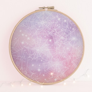 Könnte beinhalten: Ein Kreuzstich-Stickrahmen mit einem Himmelsdesign. Der Stoff zeigt einen Farbverlauf aus Rosa, Lila und Blau, mit einem weißen Spinnennetz-Muster und Sternen. Der Holzrahmen ist mit einem Messingverschluss befestigt.