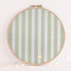 May include: A circular wooden embroidery hoop containing a piece of fabric with a vertical striped pattern in sage green and white. The fabric has a visible grid texture characteristic of Aida cloth, and the hoop features a metal tightening screw.