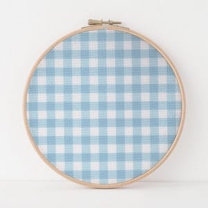 May include: A round embroidery hoop with a light blue and white gingham pattern. The fabric is stretched taut within a natural wood hoop, secured with a metal clasp. The design features a classic checkered pattern.