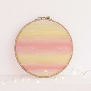 Könnte beinhalten: Ein runder Stickrahmen mit einem rosa-gelben Ombre-Design. Das Design hat einen subtilen Glitzereffekt. Der Rahmen befindet sich auf einer weißen Oberfläche mit einer Lichterkette.