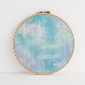 May include: A circular wooden embroidery hoop framing a piece of fabric with a printed watercolor design. The fabric features a soft, blended pattern of light blue, teal, and pale purple hues, resembling a cloudy sky or a nebula with white speckles.