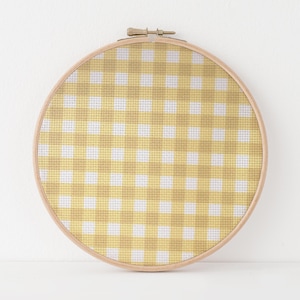 May include: A round embroidery hoop featuring a yellow and white gingham pattern. The fabric is stretched taut within a natural wood hoop, creating a decorative piece. The design is a classic, country-style pattern.
