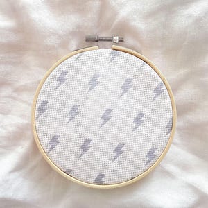 May include: A circular wooden embroidery hoop containing white fabric with a repeating pattern of small, light grey lightning bolt icons stitched in a cross-stitch style. The hoop features a metal tension screw at the top for securing the fabric.