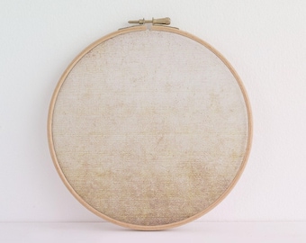 May include: A circular wooden embroidery hoop framing a piece of fabric with a gradient pattern that transitions from a light off-white at the top to a textured, mottled golden-brown hue at the bottom, featuring a metal tightening screw at the top.