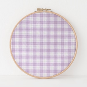 May include: A round embroidery hoop with a light wood finish holds a cross-stitch design. The design features a gingham pattern in shades of lavender and white. The fabric is stretched taut within the hoop, creating a finished piece of textile art.