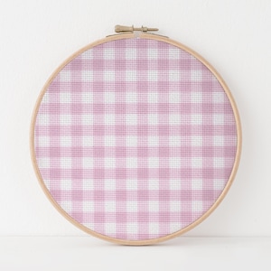 May include: A circular wooden embroidery hoop containing fabric with a light pink and white gingham check pattern. The fabric is stretched taut within the frame, which features a metal tension screw at the top for securing the material in place.