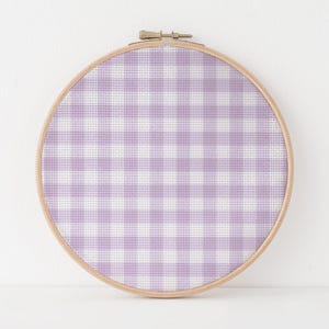 May include: A circular wooden embroidery hoop framing a piece of fabric with a light purple and white gingham check pattern. The fabric is stretched taut within the hoop, which features a metal tightening screw at the top center of the frame.