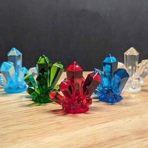 May include: Five crystal clusters in different colors: light blue, green, red, blue, and clear. The crystals are arranged on a wooden surface.