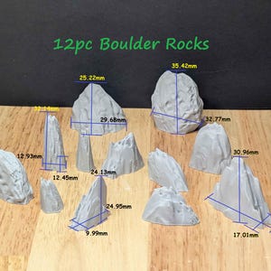May include: A set of twelve grey boulder rocks of different sizes, with measurements in millimetres. The rocks are displayed on a wooden surface, with the text "12pc Boulder Rocks" in green at the top.