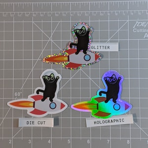 May include: Three black cat stickers on a rocket ship. The stickers are available in three finishes: glitter, die cut, and holographic.