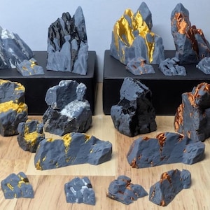 May include: A collection of gray, gold, and copper-colored miniature rock formations. Some are displayed on black platforms. The rocks vary in size and shape, with textured surfaces and metallic accents. The overall theme is geological or decorative.
