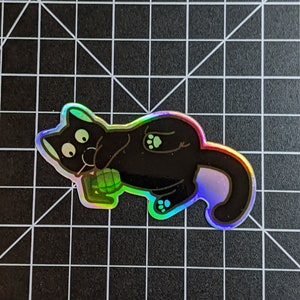 May include: A black cat sticker with iridescent holographic foil. The cat is lying on its side with its paws in the air and its eyes wide open. The cat is holding a green object in its mouth.