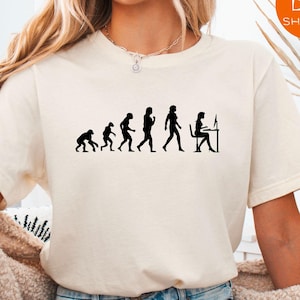 May include: A white t-shirt with a black silhouette of the evolution of humans ending with a woman sitting at a computer. The text "Next Day Shipping" is in the top right corner of the image.