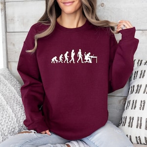 May include: Burgundy crewneck sweatshirt featuring a white graphic of the evolution of man, ending with a person at a computer. The sweatshirt is made of a soft material.