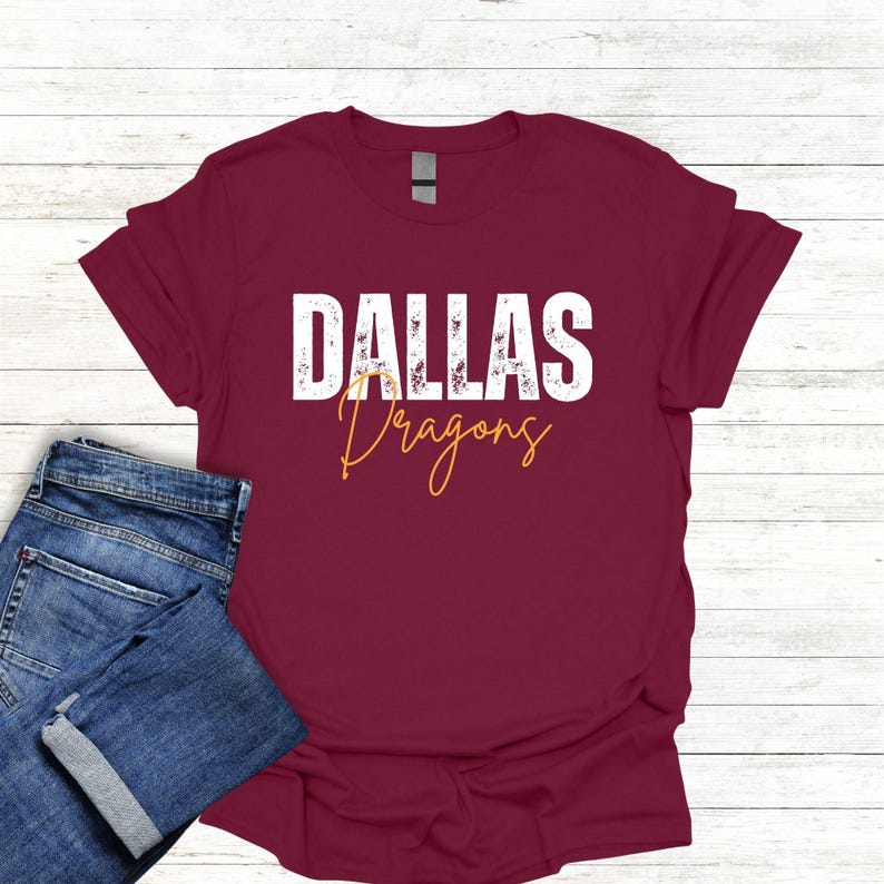 K&ouml;nnte beinhalten: Burgunderfarbenes T-Shirt mit wei&szlig;em Text, der "Dallas Dragons" in einer verwitterten Schriftart lautet. Der Text "Dragons" ist in Gelb-Orange.