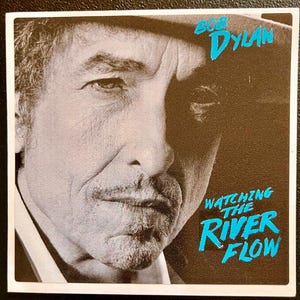 May include: Square album cover featuring a black and white portrait of Bob Dylan. The text "BOB DYLAN" is in turquoise script at the top, with "WATCHING THE RIVER FLOW" in turquoise script below.