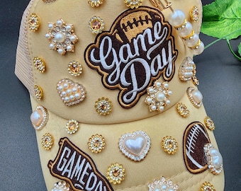 Może przedstawiać: Beżowa czapka z daszkiem z siatkowym tyłem, ozdobiona perłami i dżetami. Czapka zawiera napis "Game Day" w brązowo-białym haftowanym wzorze, a także naszywki z motywem futbolu amerykańskiego.